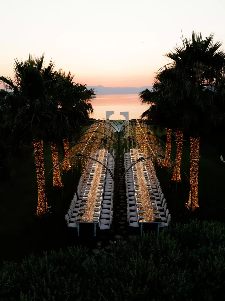 Long reception tables under glowing lights and palm trees at Sea View by En Kipo during sunset
