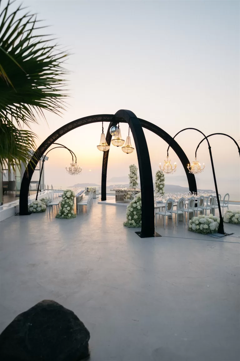Outdoor wedding reception setup with chandeliers and long tables overlooking sunset sea views