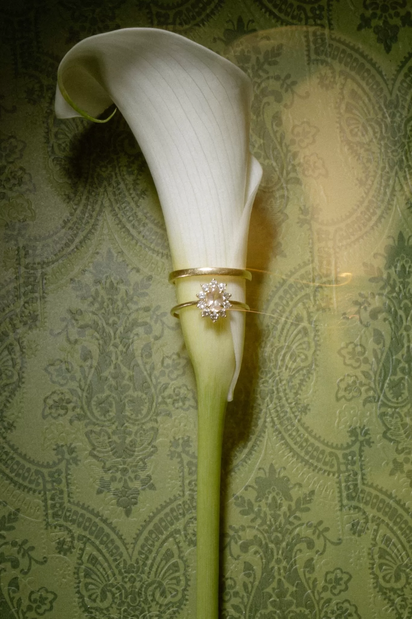 Close-up of a diamond engagement ring placed on a white calla lily against a soft green patterned background.