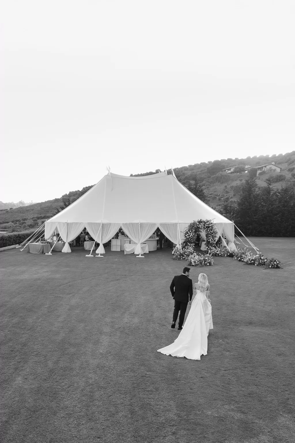 Bride and groom walking across the lawn toward an elegant sailcloth wedding tent decorated with flowers.