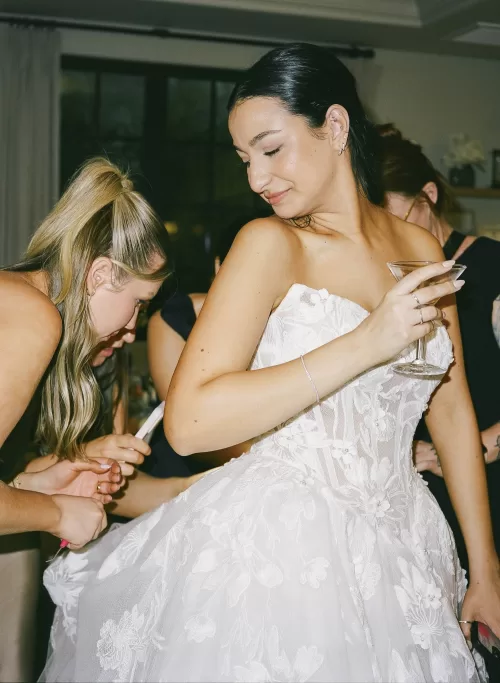 Bride smiles with a cocktail while her dress is adjusted during the reception