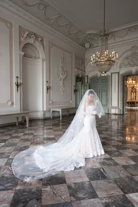 Bride standing alone with a long flowing veil in a grand historic ballroom with chandeliers