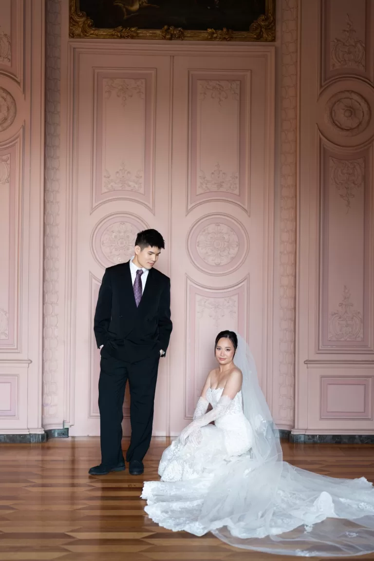 Bride seated in a soft pink ornate room while groom stands nearby in a refined portrait scene