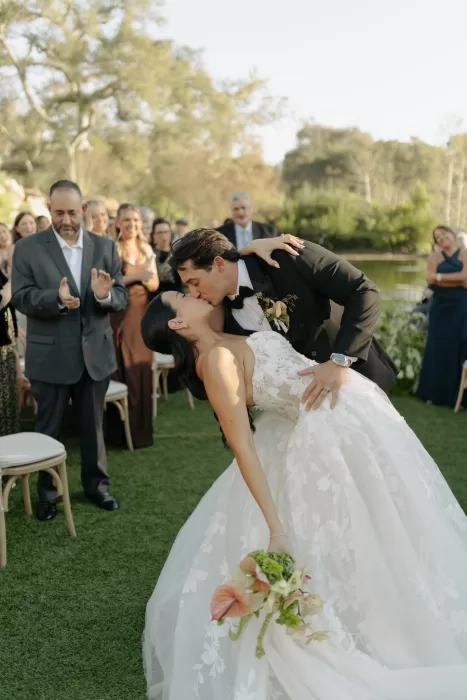 Couple share first kiss at ceremony as guests applaud beside lakeside setting
