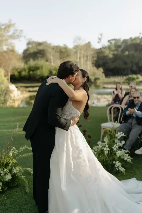 Couple embrace and kiss at altar framed by florals and calm lakeside backdrop