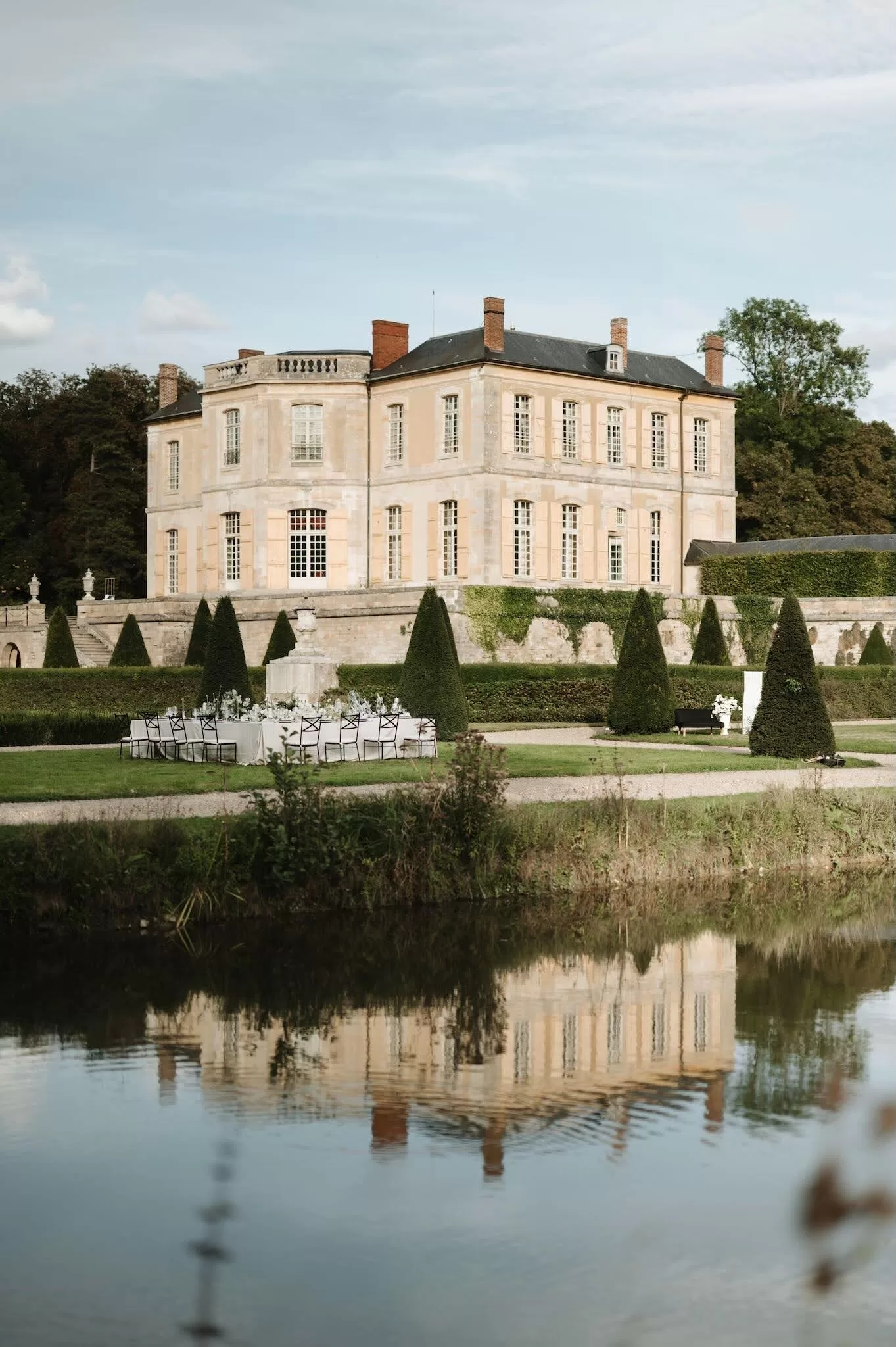 Outdoor dining table set on the lawn beside the reflecting pond at Château de Villette, surrounded by formal gardens.