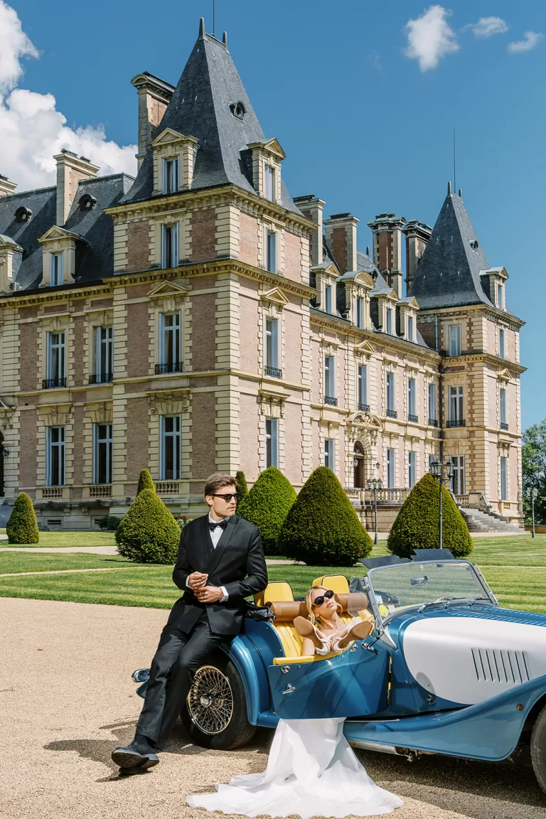 Couple pose with a blue vintage car outside the château gardens