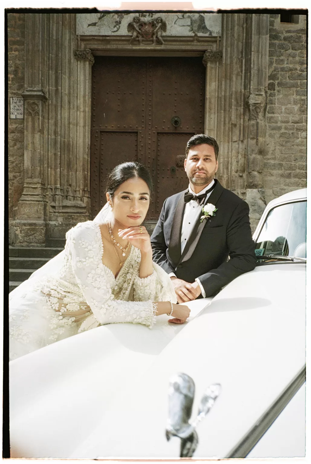 Bride and groom posing beside vintage car outside historic Barcelona church doorway