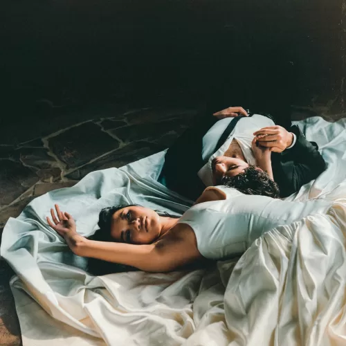 The couple lie across the bride’s gown in a dramatic, softly lit portrait.