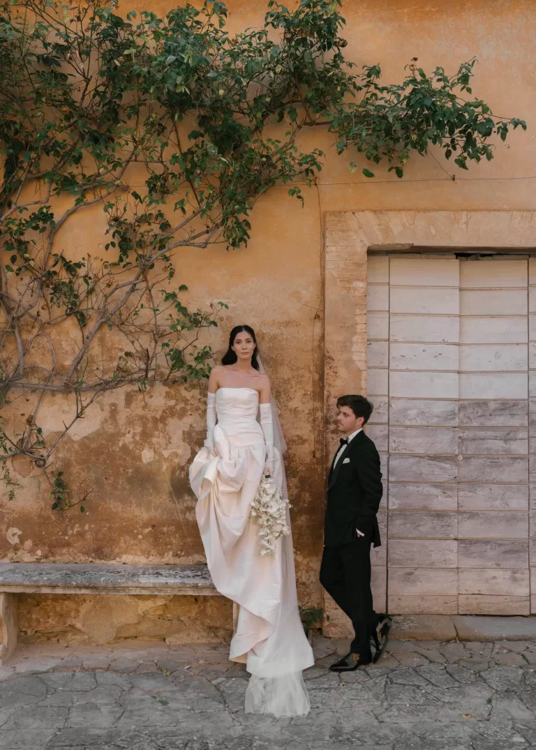 Bride and groom posing against warm textured wall with greenery in romantic Italian setting