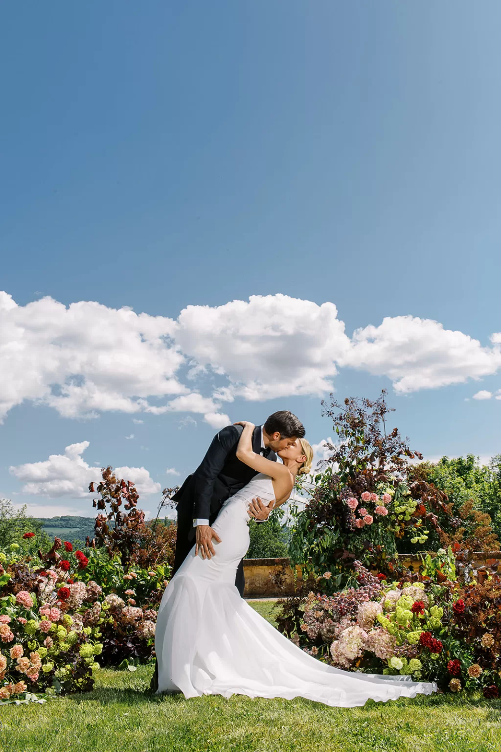 Couple share a kiss among lush garden florals beneath a wide blue sky