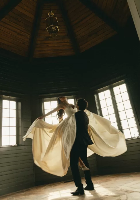 The groom lifts the bride as her gown moves through the warm window light.