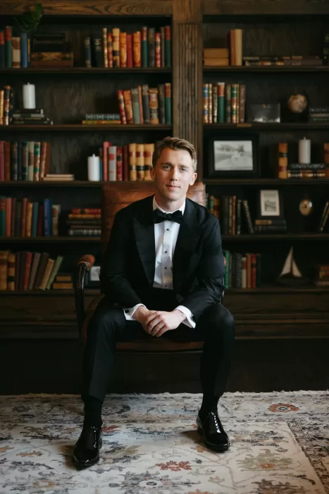 Groom portrait in a library setting at The Manor House, wearing a classic black tuxedo.