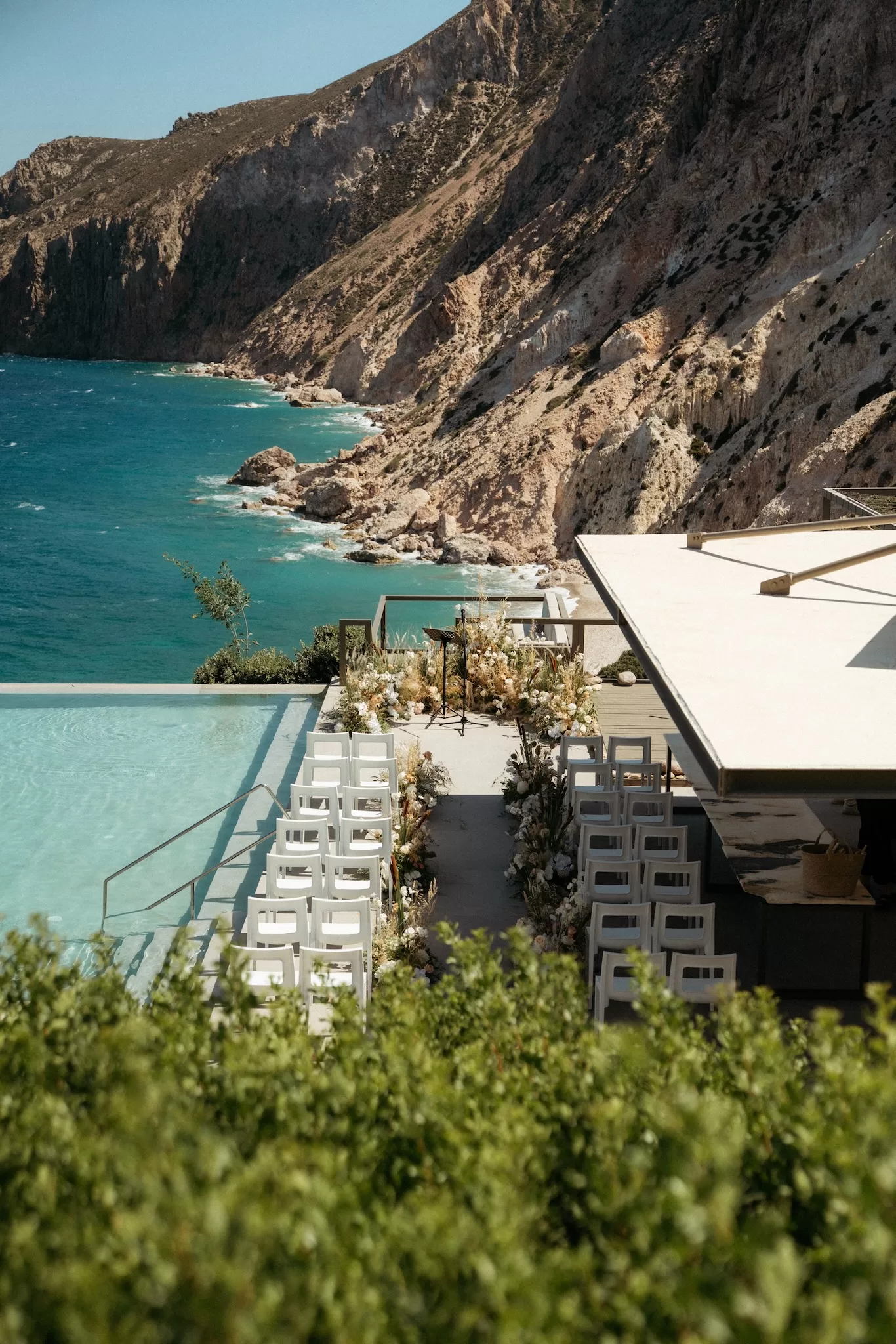 Poolside wedding ceremony layout at Milos Cove overlooking turquoise sea and rugged cliffs.