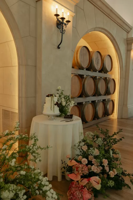 Wedding cake displayed beside wooden barrels and soft floral arrangements inside Monserate Winery