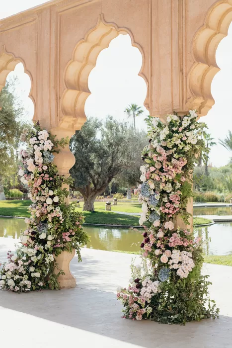 Floral ceremony pillars overflowing with soft pink and white blooms beside the water at Palais Namaskar