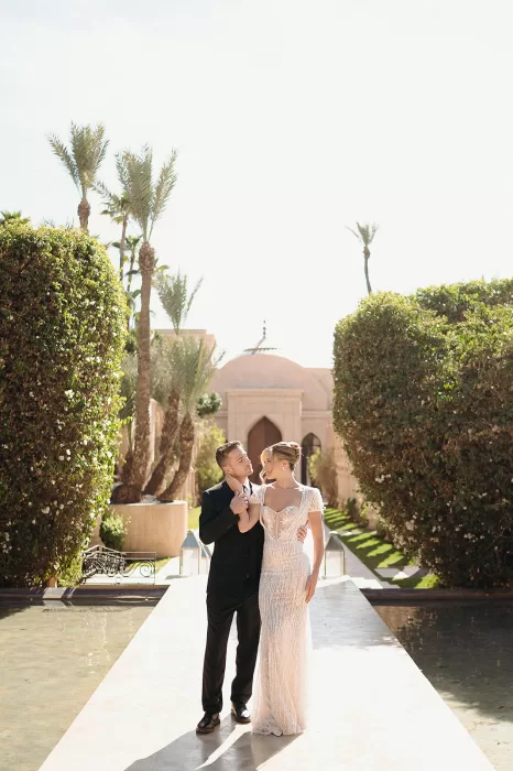 Bride and groom embracing on garden walkway lined with palms and water at Palais Namaskar