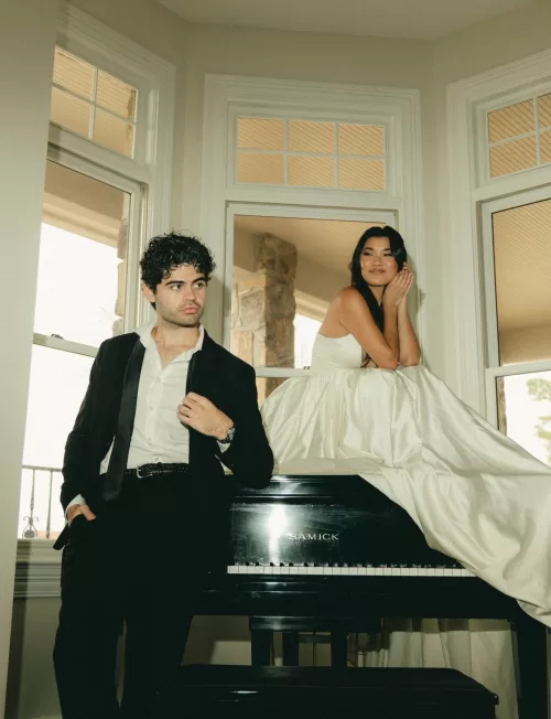 The couple pose with a grand piano beneath warm light and tall windows.