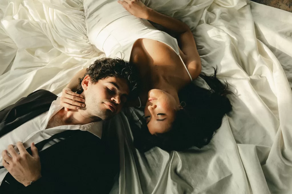 The couple lie together in warm sunlight, surrounded by soft folds of fabric.