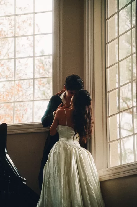 Quiet portrait of the couple embracing beneath tall windows in warm natural light.