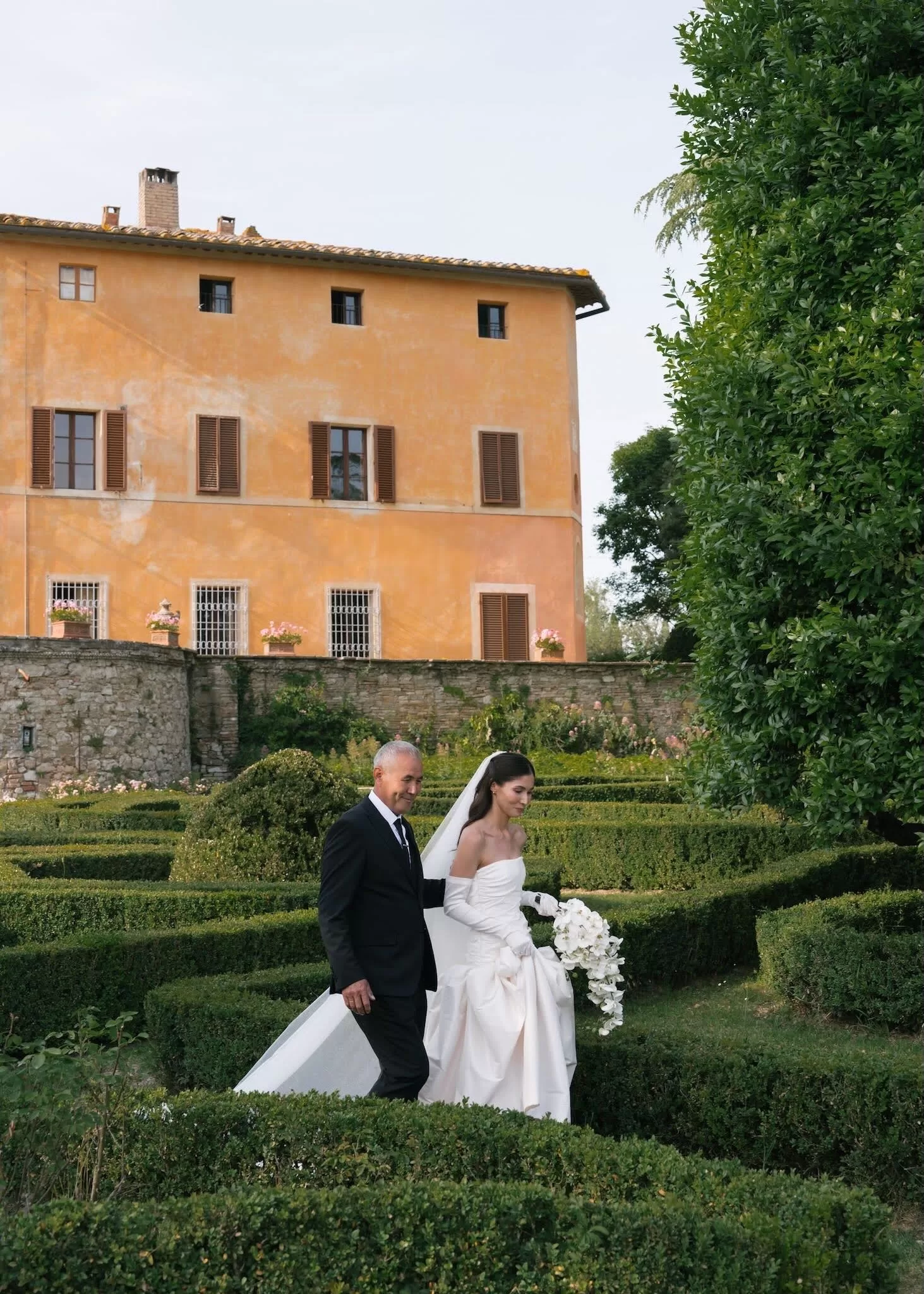 Bride walks through the gardens of Villa Catignano on her father’s arm, framed by box hedges and the villa’s ochre façade.