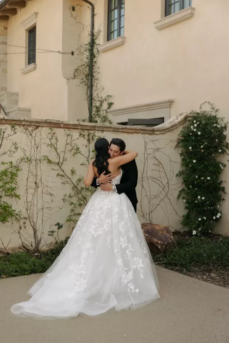 Bride and groom share a quiet embrace against a sunlit courtyard wall with climbing greenery