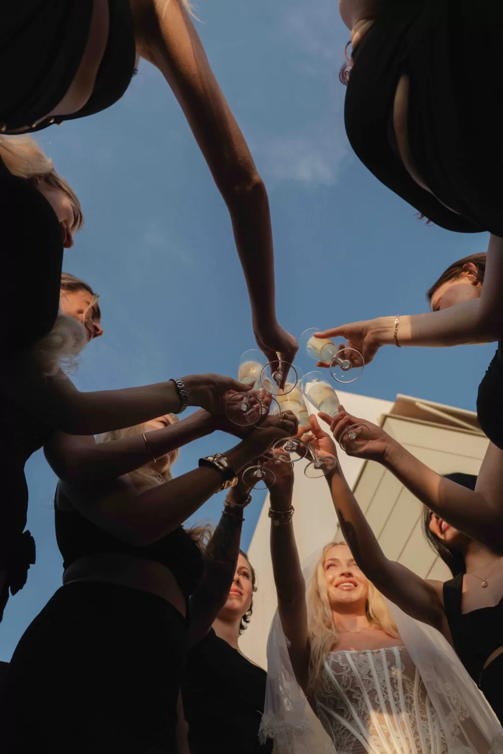 Bride and bridesmaids in black dresses clinking champagne glasses at sunset during a chic hen party celebration.