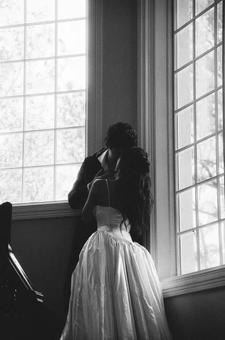 Black and white portrait of the couple kissing beside tall, light-filled windows.