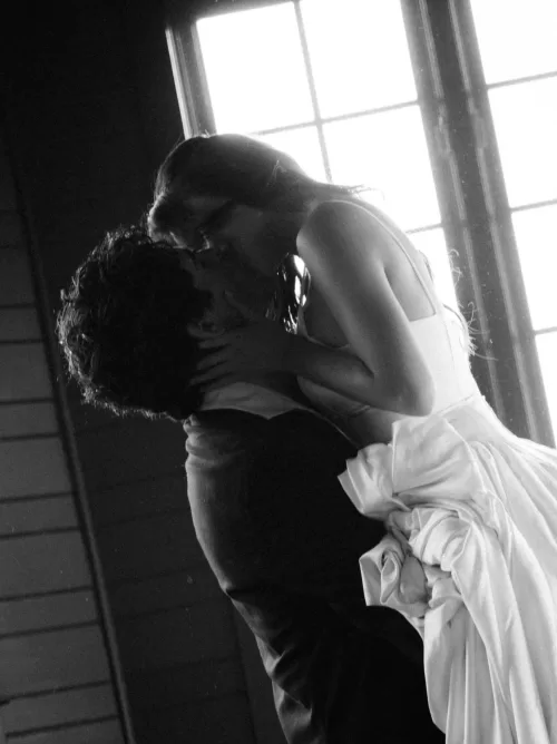 Black and white silhouette of the couple kissing beside a bright window.