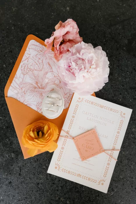 Coral wedding stationery, rings, and soft florals styled against dark stone at Acre Baja.