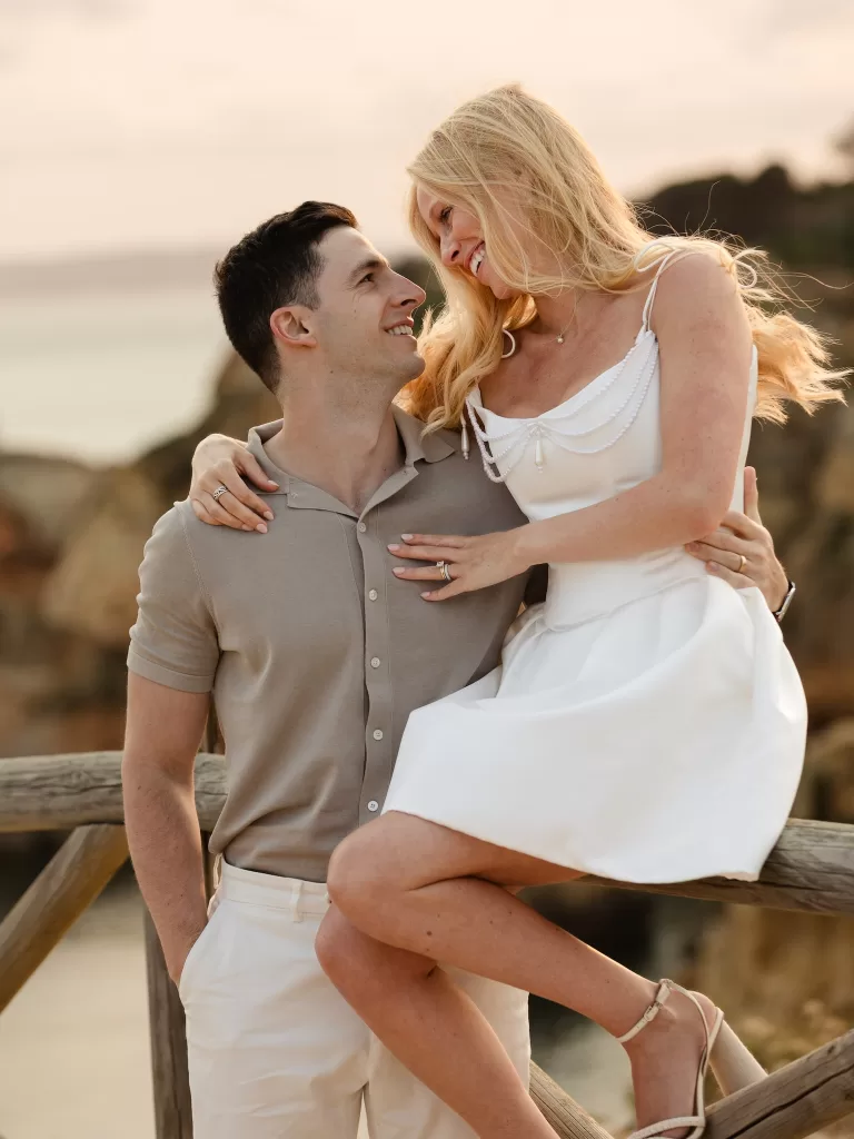 Relaxed couple portrait on a wooden fence with soft Algarve coastline behind them.
