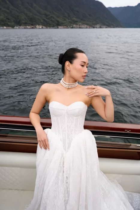 Bride posed on a lake boat in a lace gown, with dark water behind her.