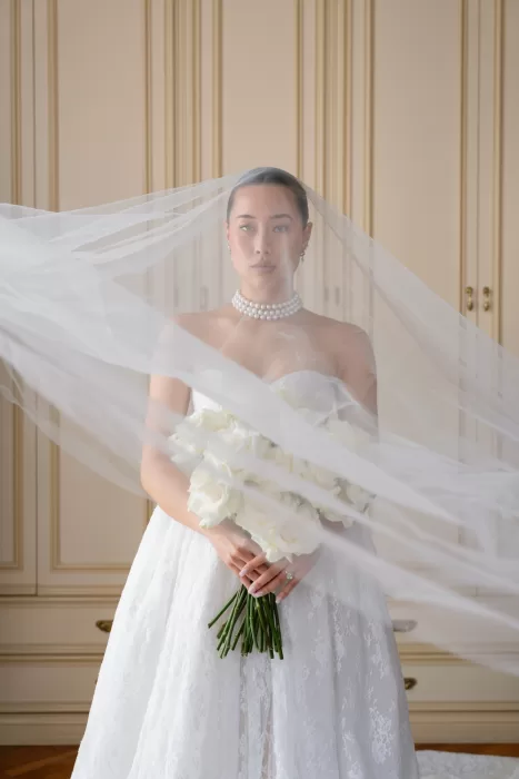 Bride holding white roses beneath a sheer veil in a softly lit villa room.