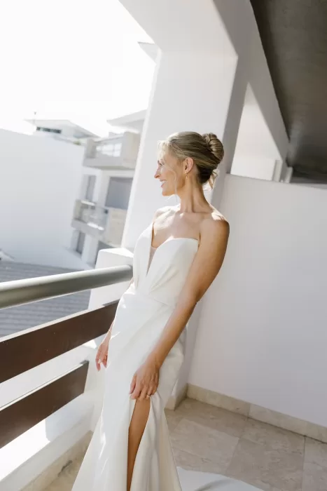 Bride in a sleek gown smiling on a sunlit balcony before the ceremony.