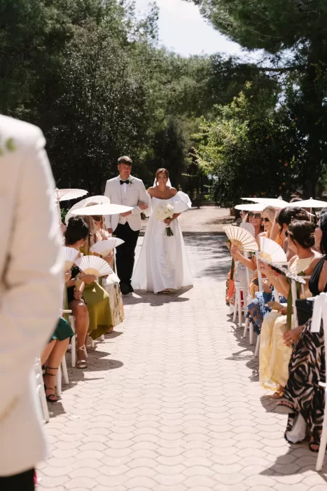 Bride walking down aisle with guests holding fans, bright Mediterranean sun overhead