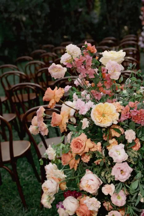 Soft peach and pink ceremony flowers arranged beside rows of wooden chairs.