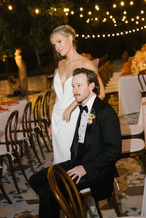 Bride and groom seated together beneath string lights before the outdoor reception.