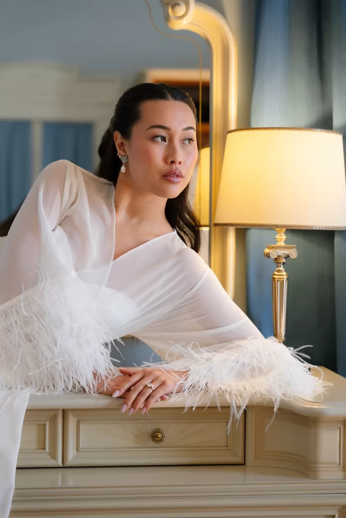 Bride leaning beside a glowing lamp in a feathered robe and pearl earrings.