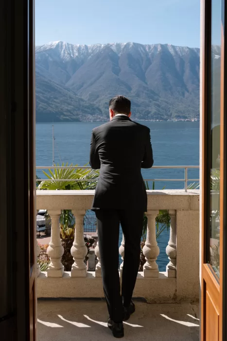 Groom looking across Lake Como from a sunlit balcony with mountains beyond.
