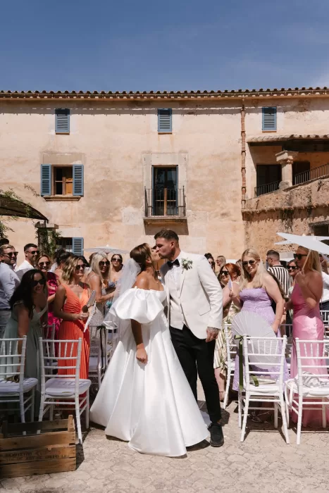 Bride and groom kiss in a sunlit Mallorca finca courtyard surrounded by guests after the ceremony