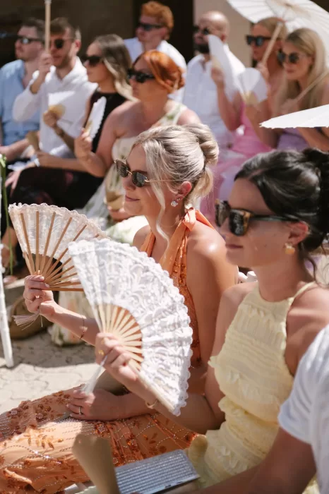Wedding guests seated outdoors using handheld fans during a sunny ceremony at a Mallorca finca