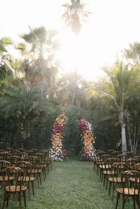 Sunlit ceremony chairs face colourful floral pillars beneath towering palms.