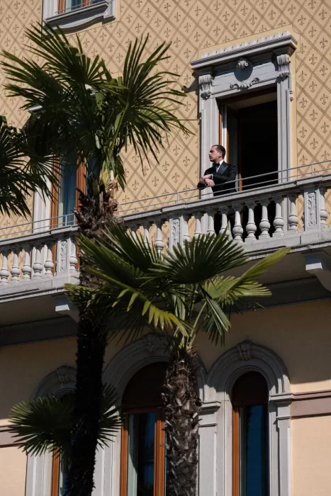 Groom in a black tuxedo standing on a patterned villa balcony framed by palms.