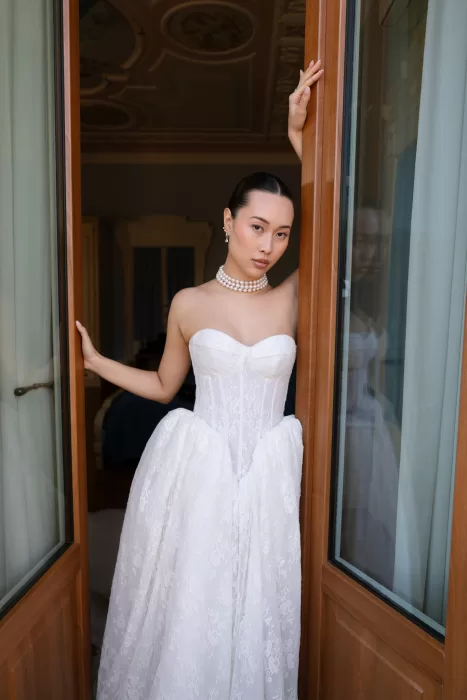 Bride framed in open wooden doors, wearing lace and pearls in a villa suite.
