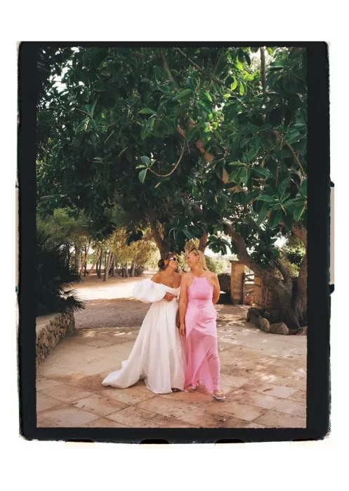 Bride and guest strolling under trees, relaxed moment in shaded garden pathway