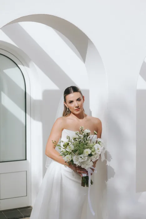 The bride holds a soft white bouquet beneath clean white arches and sharp sunlight.