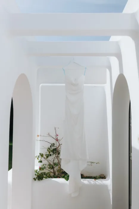 A white wedding dress hangs quietly inside a sunlit arched corridor.