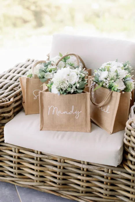Personalised jute bags filled with white floral arrangements on a woven chair.