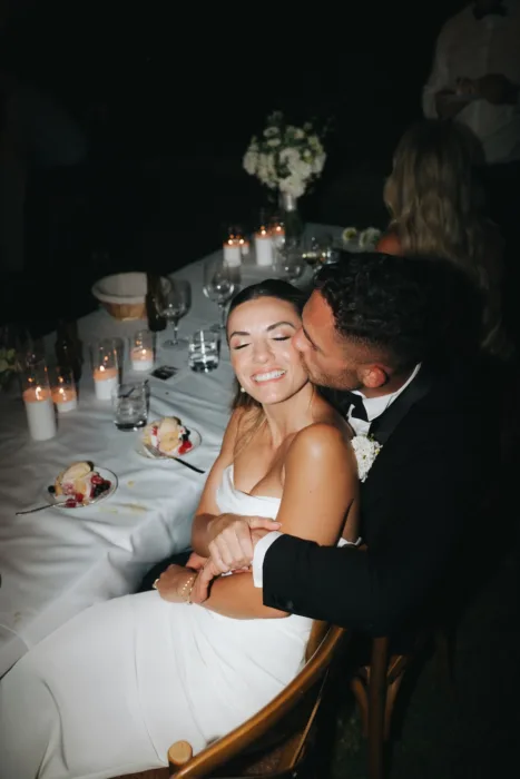 The groom kisses the bride during a candlelit outdoor dinner with glasses and dessert plates.
