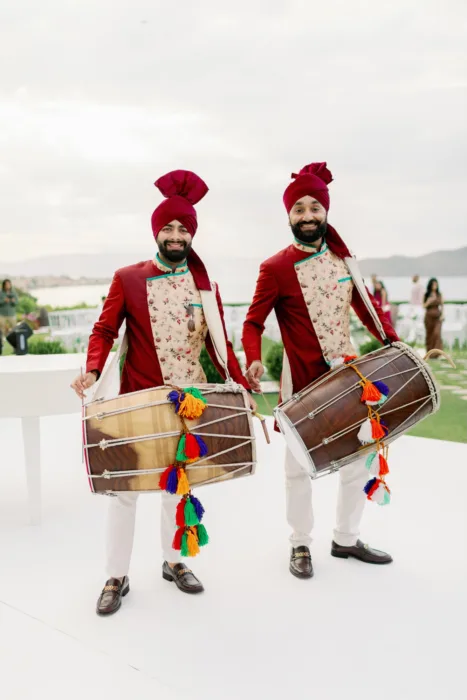 Dhol drummers in red outfits perform on the terrace with the coast behind them.
